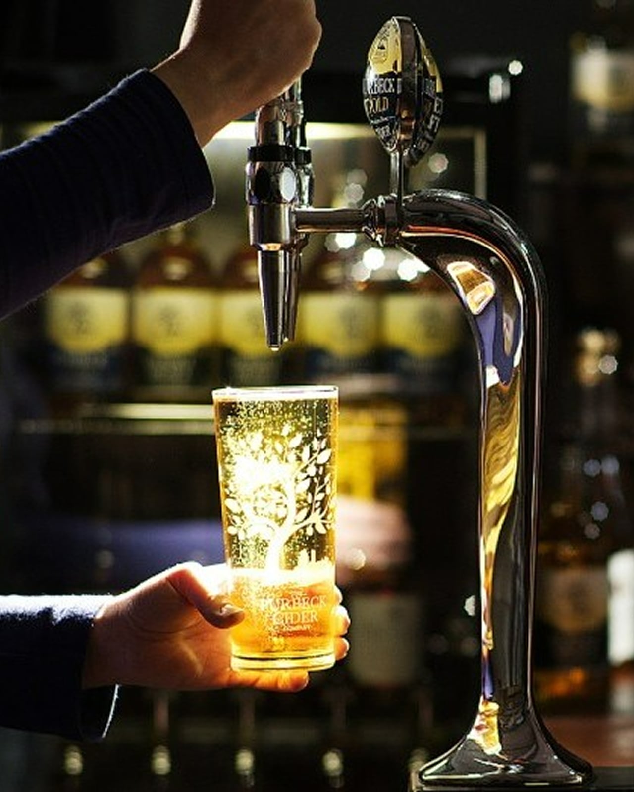 Person pouring beer from a tap into a glass with blurred bottles in the background